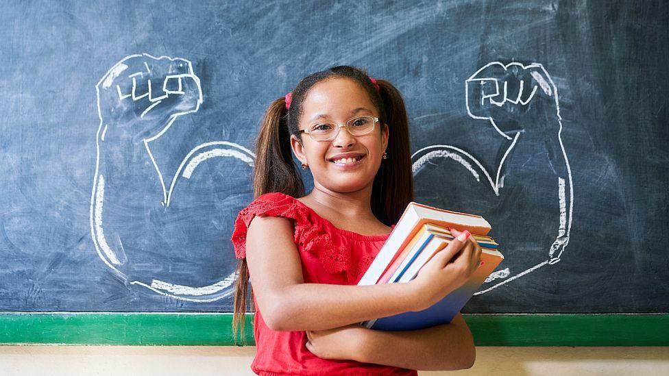 Niña en clase frente a un pizarrón en el que están dibujados a cada lado de ella brazos musulosos