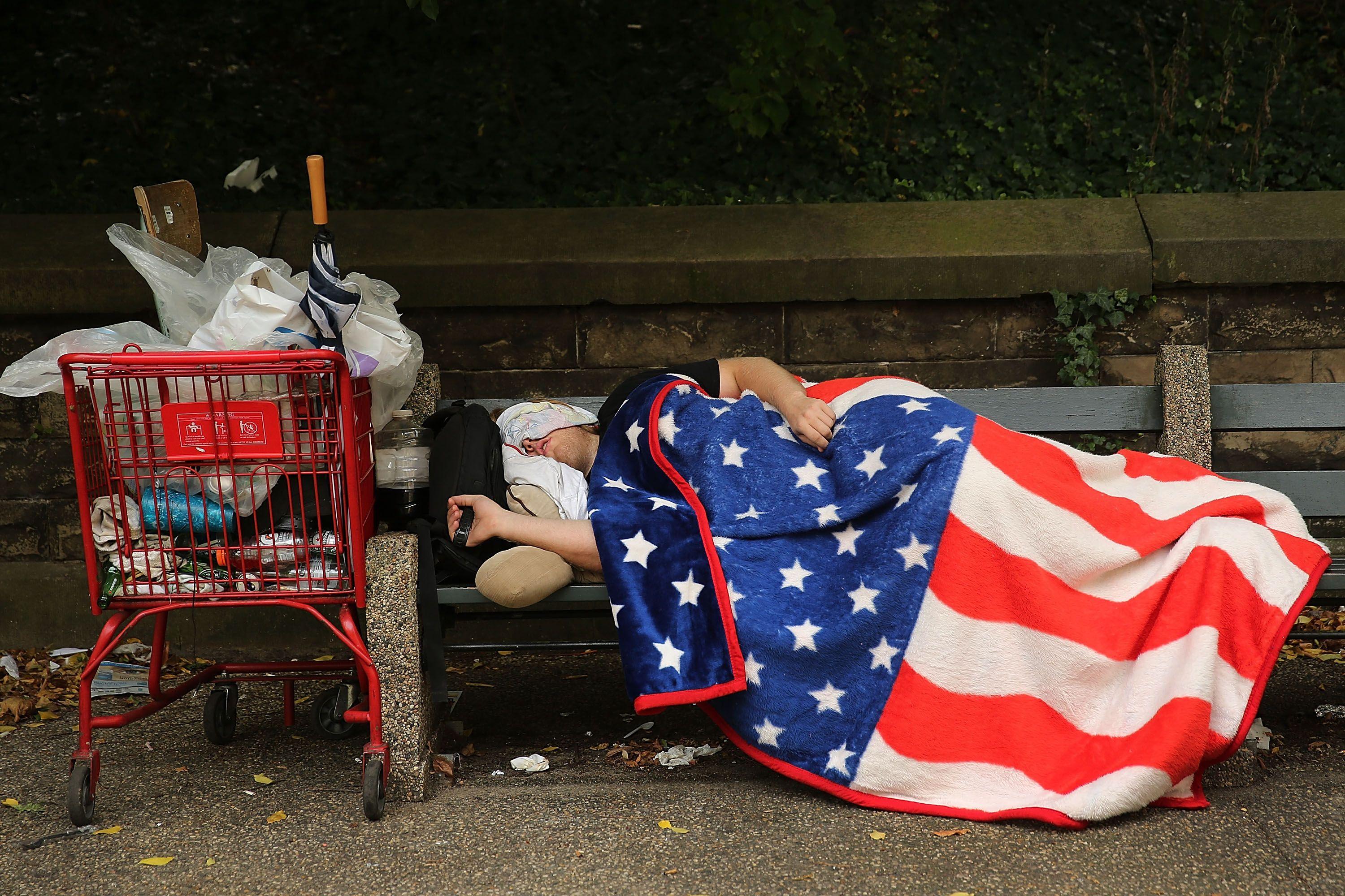 Una persona duerme en un banco callejero en Nueva York envuelta en una colcha con el diseño de la bandera de EE.UU.