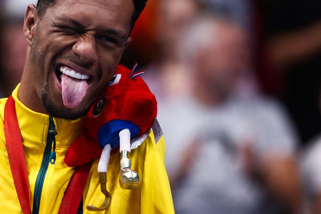 Gabriel Araújo, de Brasil, celebra su medalla de oro en natación con la lengua afuera y sosteniendo la mascota de los Juegos con su cabeza y hombro.