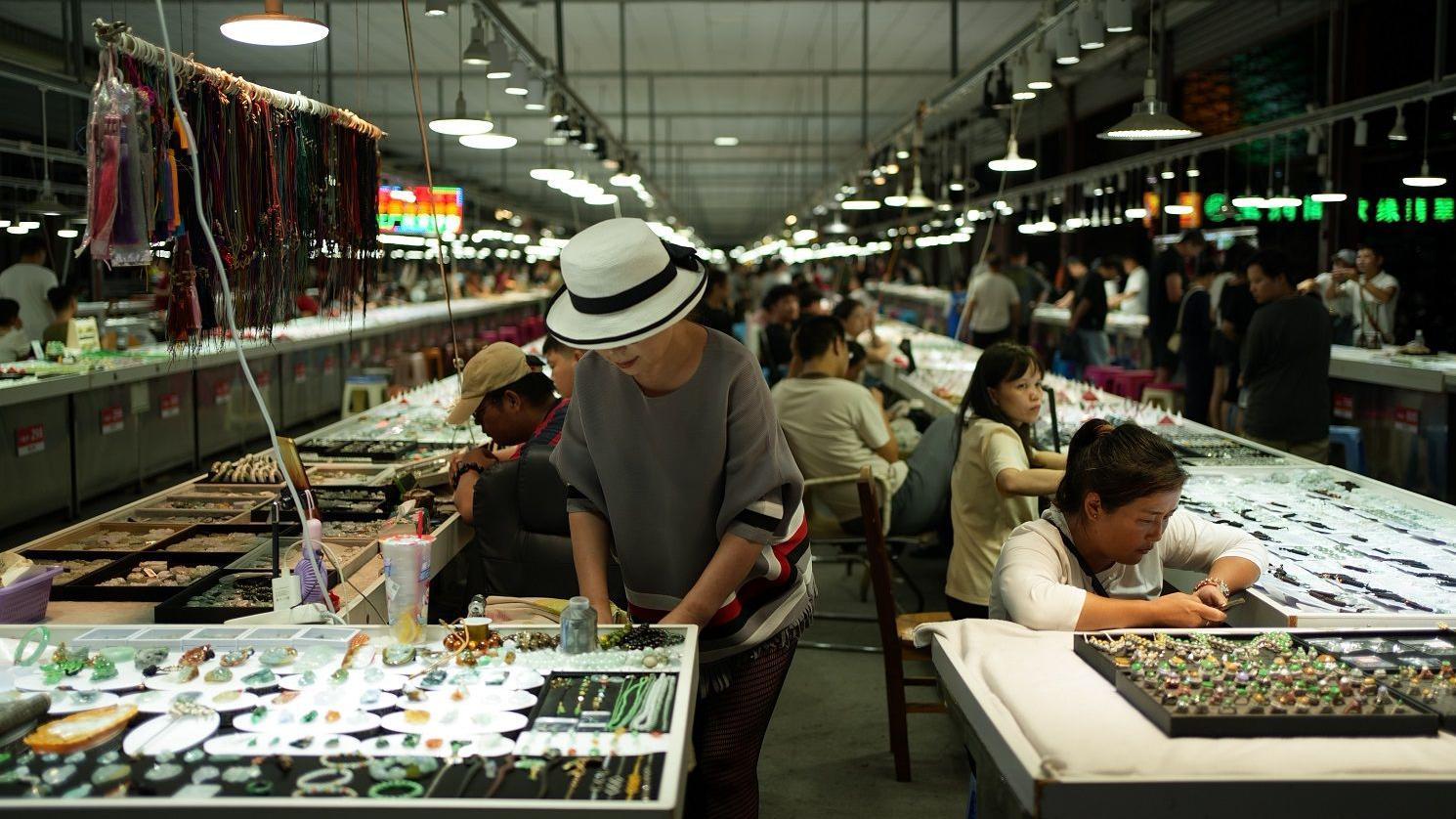 En el mercado de Ruili, las mujeres se sientan y permanecen de pie junto a los puestos de venta de joyas.