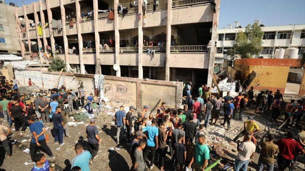 Gente alrededor de una escuela semi derruida a causa de los bombardeos. 