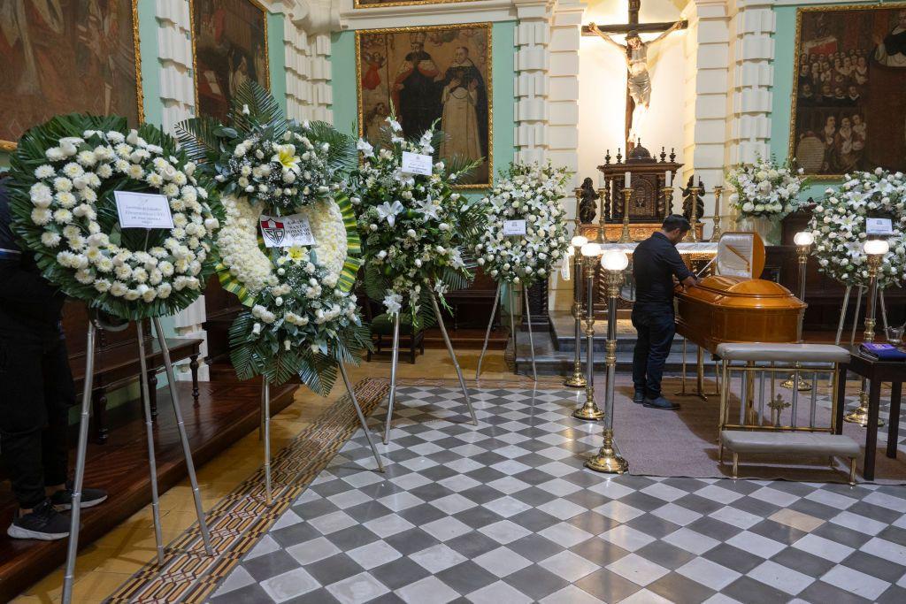 Un hombre rinde homenaje al fallecido sacerdote e intelectual peruano Gustavo Gutiérrez en una iglesia. El ataúd está rodeado de coronas de flores blancas.