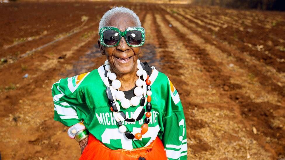 Margret Chola con una camiseta de fútbol americano, gafas, collares y una falda de colores brillantes