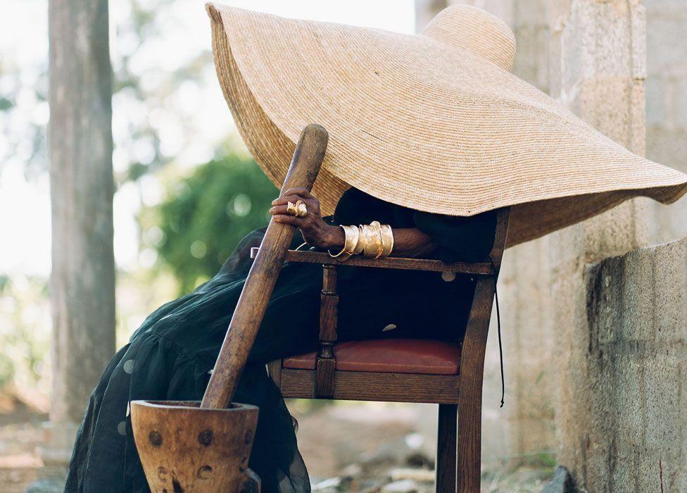 Margaret con un enorme sombrero alado que le cubre el rostro senta en una silla sosteniendo un ibende o palo para machacar cultivos