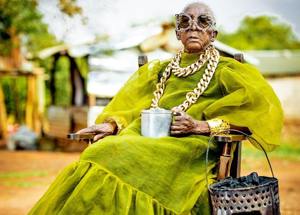 Margret Chola con un vestido con una capa de gasa, gafas, un brasero para cocinar y una taza de metal con té