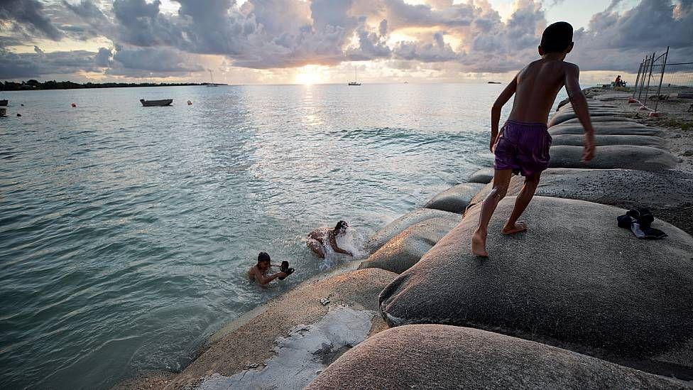 Niños jugando en una laguna y sobre bolsas de arena que buscan frenar el avance del mar