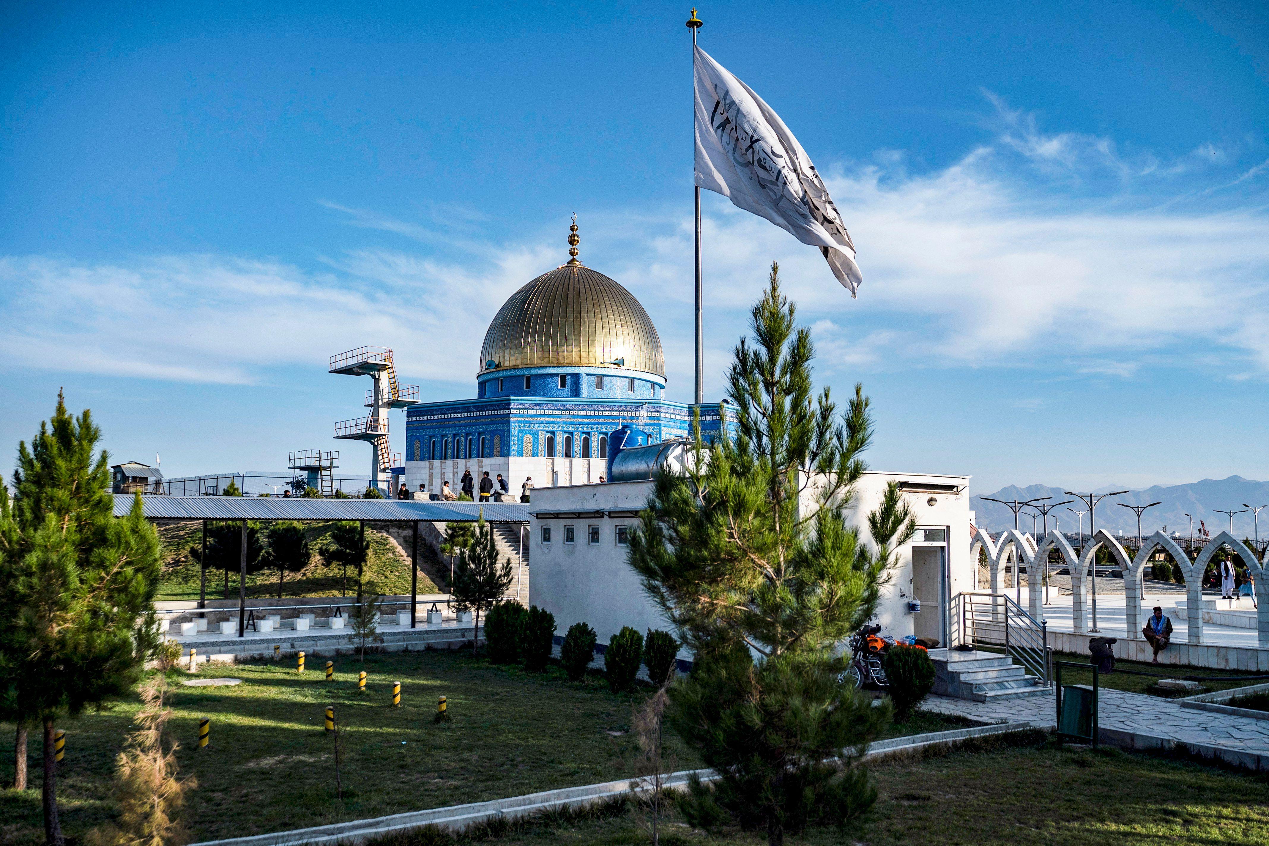 Una mezquita azul con cúpula dorada detrás de una enorme bandera ondeando en un mástil.