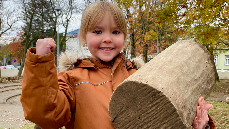 Una niña sonriente carga un tronco mientras que, con su otro brazo, hace señal de "soy fuerte!".