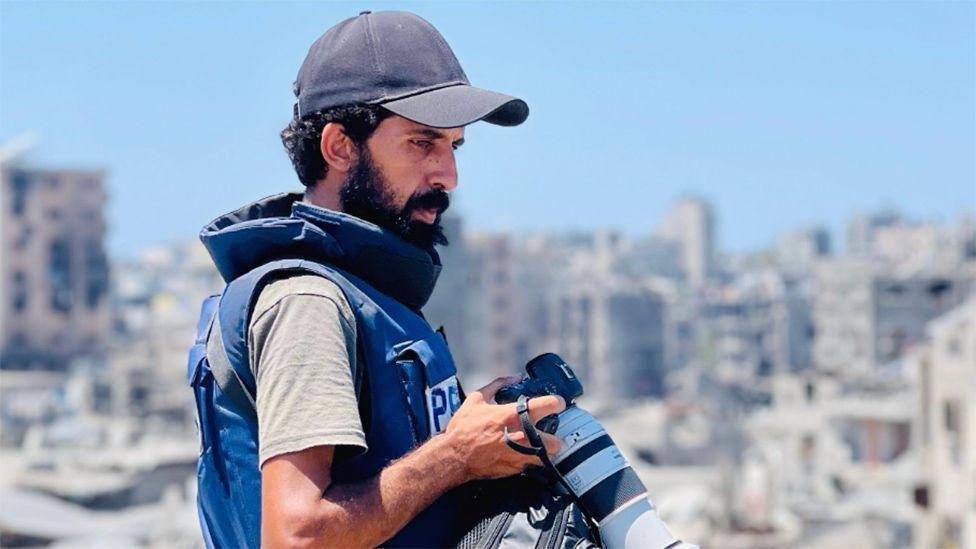 Un hombre con barba y gorra de béisbol sostiene una cámara . Está de perfil y lleva un chaleco antibalas con la palabra "Prensa". Al fondo se ve una imagen borrosa de lo que parece ser una ciudad fuertemente bombardeada.
